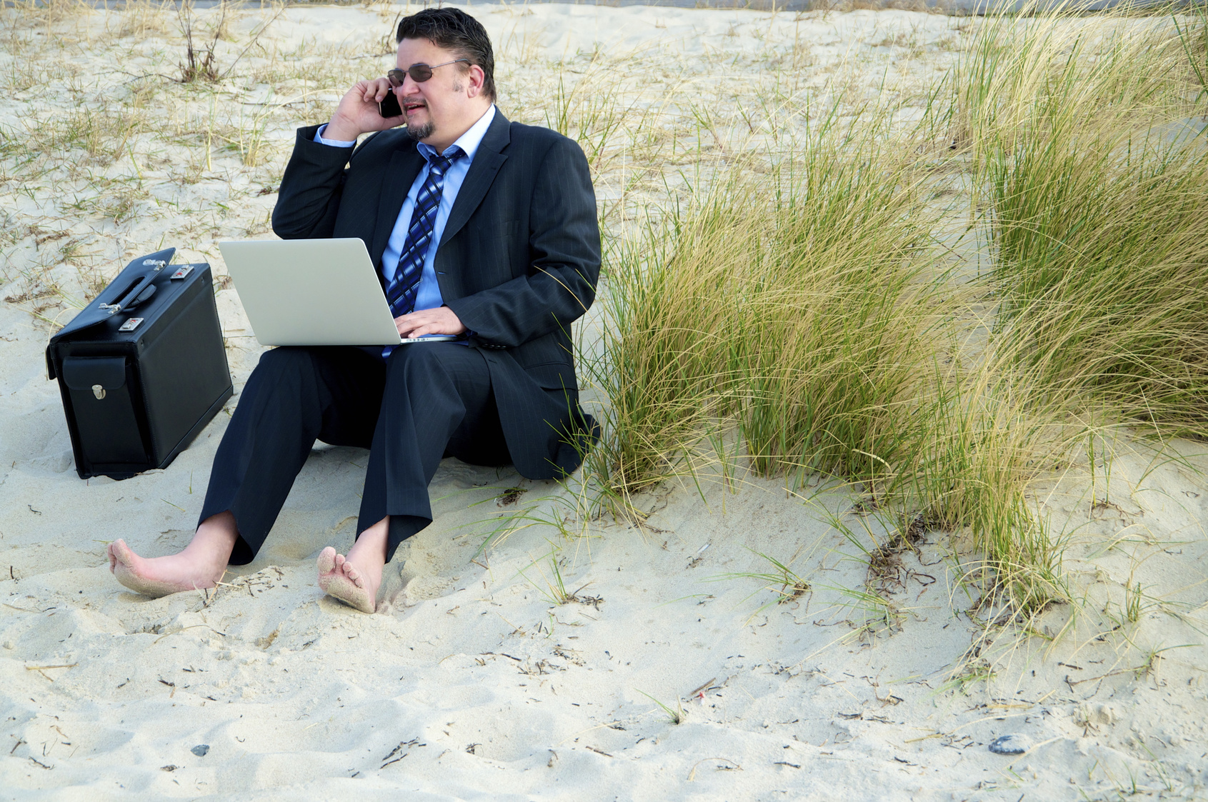 Dicklicher Geschäftsmann sitzt im Anzug am Strand, Aktentasche neben sich, Laptop auf den Beinen und das Handy am Ohr; Kurtz Detektei Hannover, Wirtschaftsdetektei Hannover, Detektiv Hannover, Detektivagentur Hannover