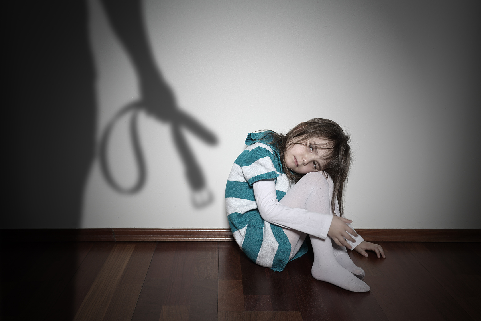 Mädchen sitzt mit angezogenen Knien auf dem Fußboden und guckt traurig. An der Wand der Schatten eines Mannes mit Gürtel in der Hand; Gewalt gegen Kinder, Kurtz Detektei Hannover, Privatdetektiv Hannover, Privatermittler Hannover, Privatdetektei Hannover